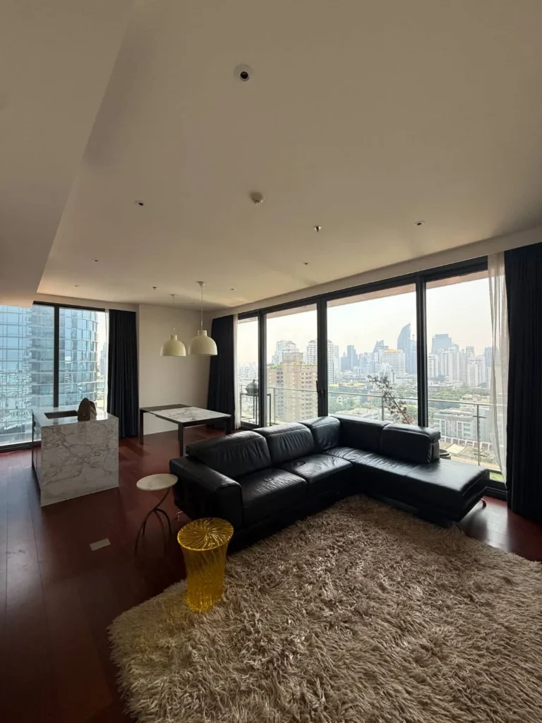 Living room with a black leather L-shaped sofa, floor-to-ceiling windows and city views, kitchen island and dining table behind